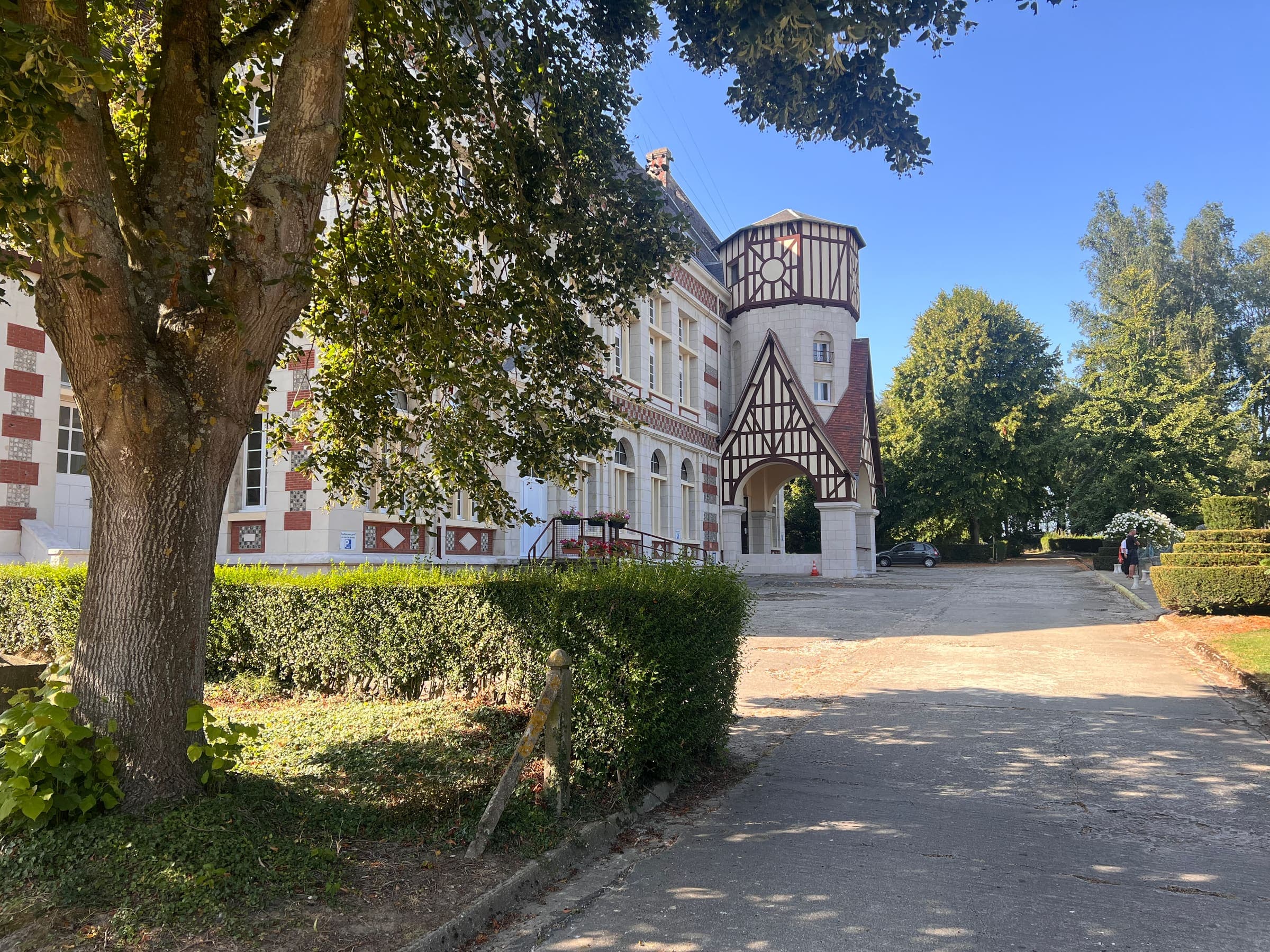 La cour devant l’église, à l’ombre d’un grand arbre, et le porche à colombages au fond.